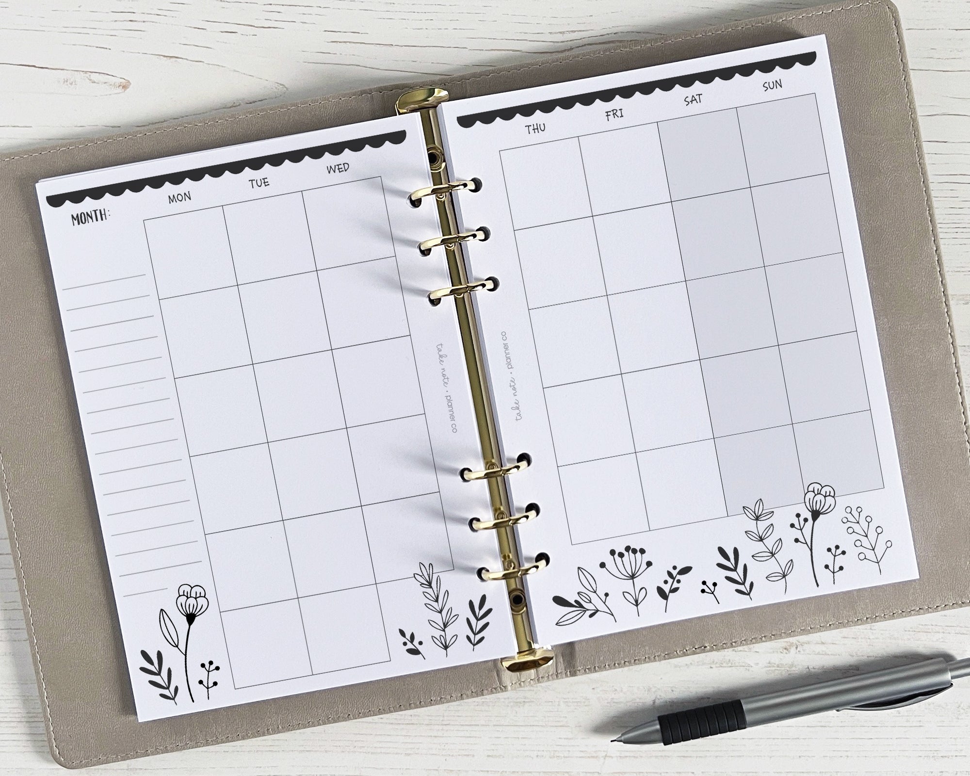A5 monthly planner inserts with a monochrome floral design, undated, displayed open in a binder with a pen beside it.