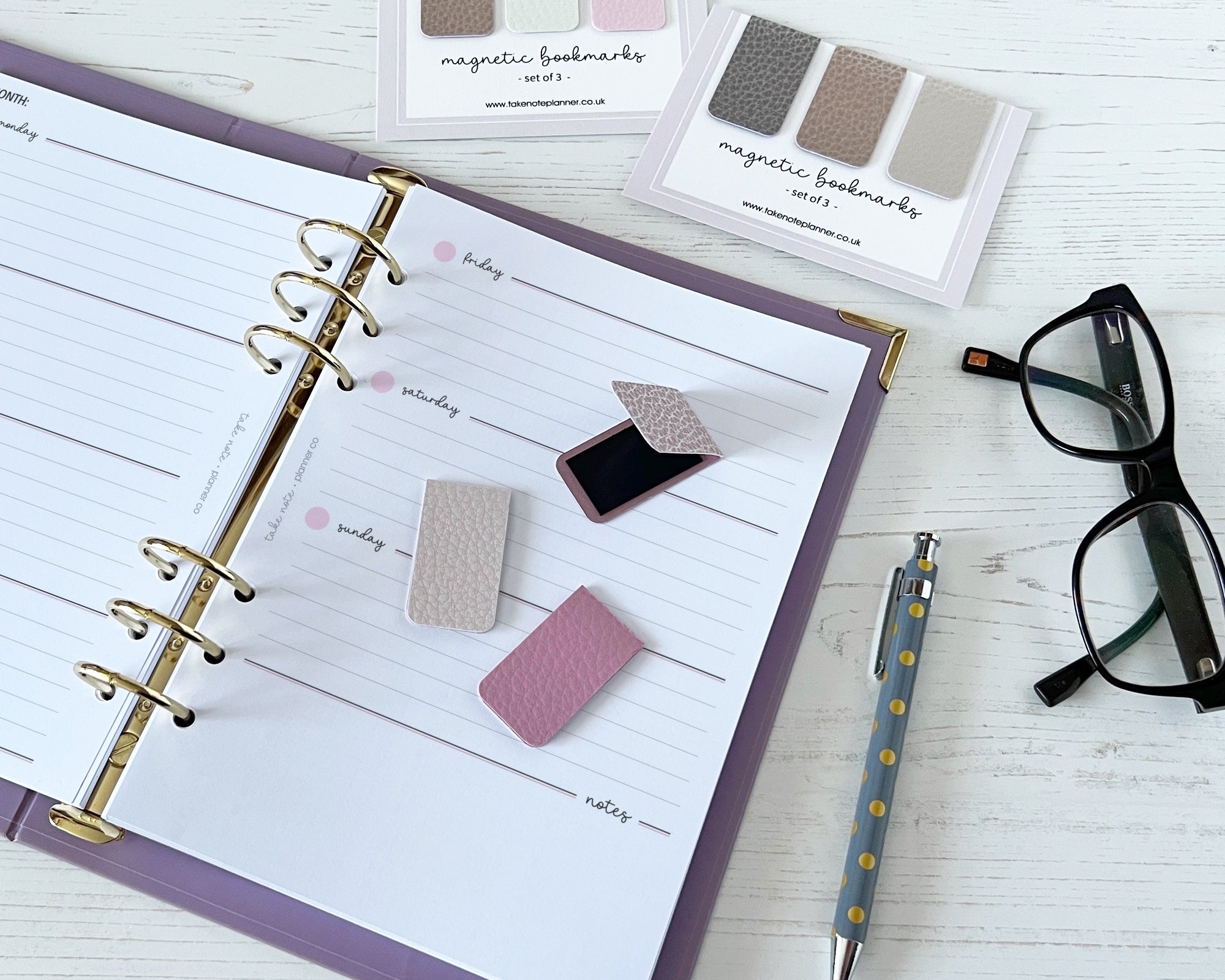 Open planner with magnetic bookmarks, pen, and glasses on a white surface
