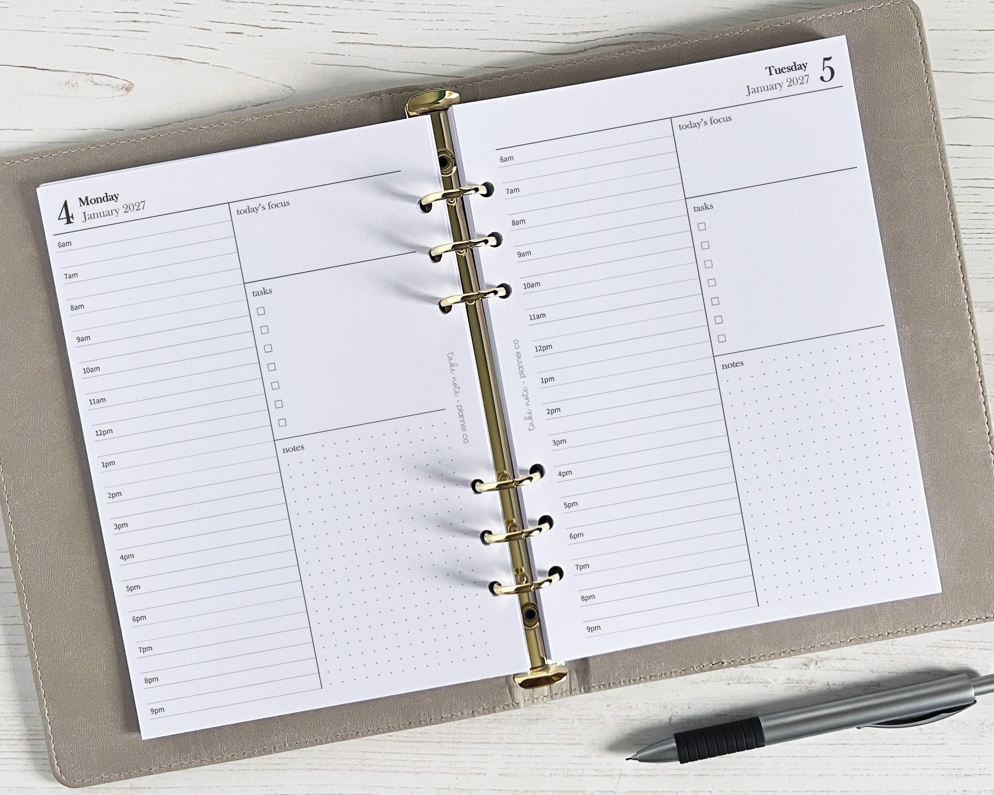 A5 daily planner inserts with a simple, elegant, minimal design, featuring one day per page with half/hourly breakdown, priorities/events box, to do list, and space for notes. The image shows the planner open to a daily insert with a pen lying next to it, on a wooden surface.