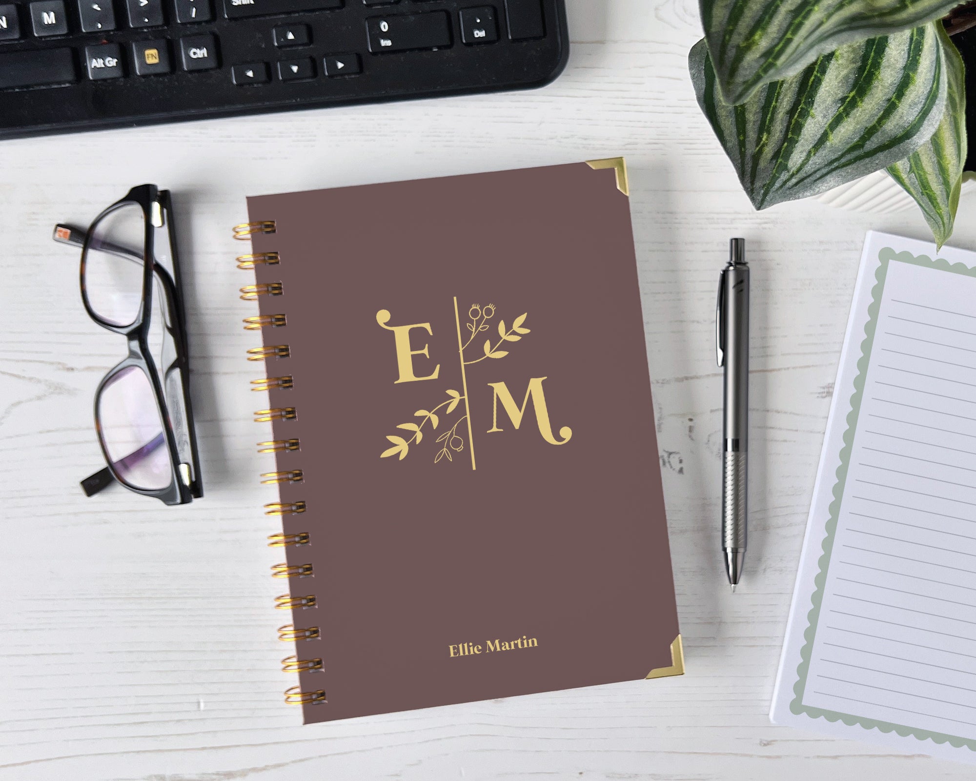 Brown notebook with gold monogram on a desk with glasses, pen, and keyboard.