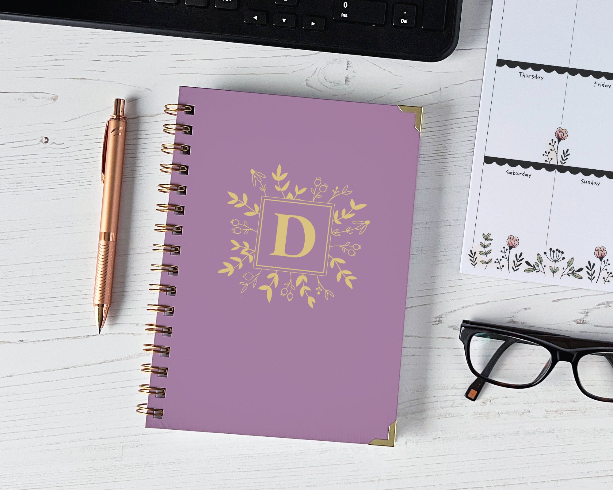 lavender notebook with gold monogram on a white surface with a pen, glasses, and calendar.