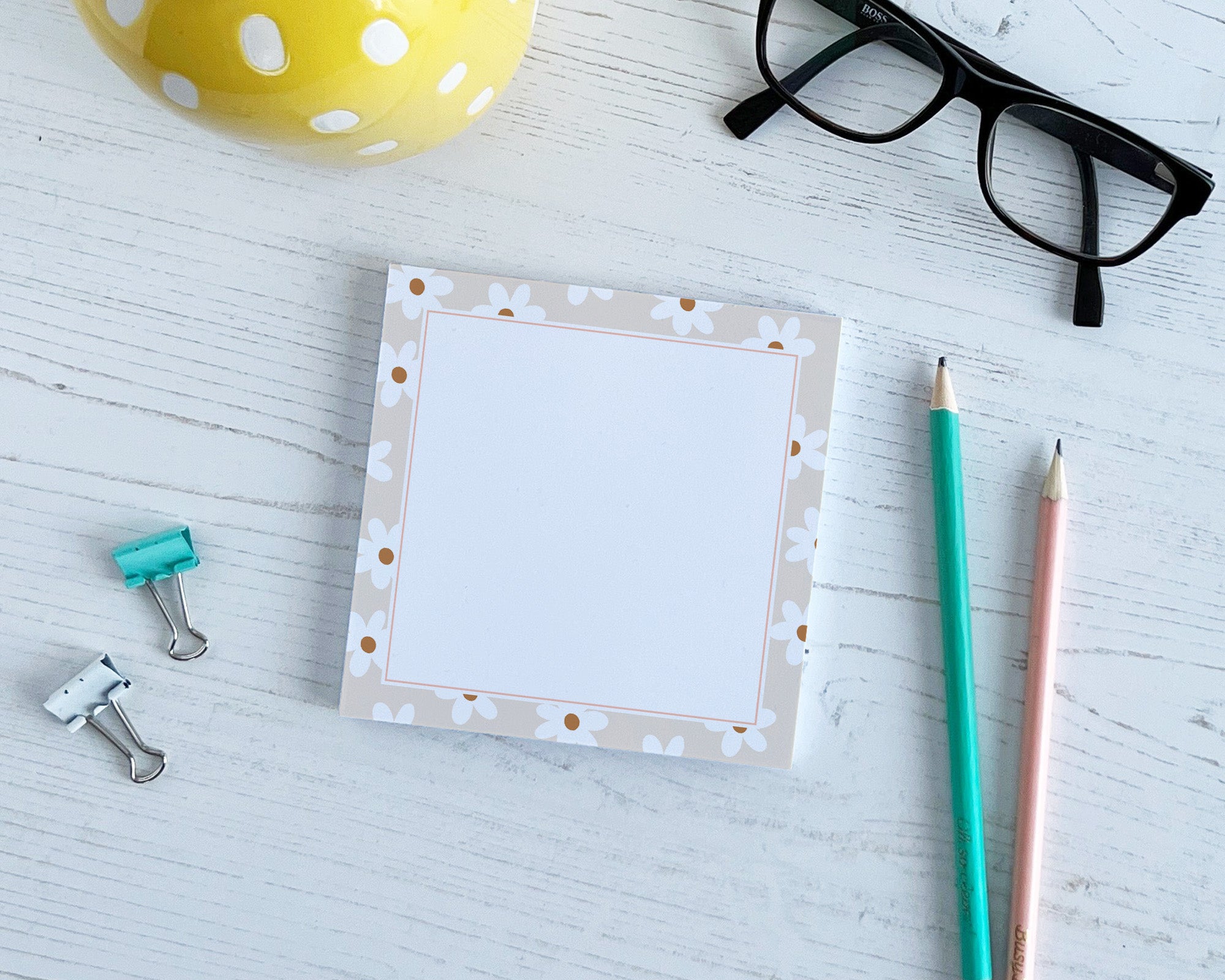 Note pad with daisy border on a light wooden surface with stationery items.