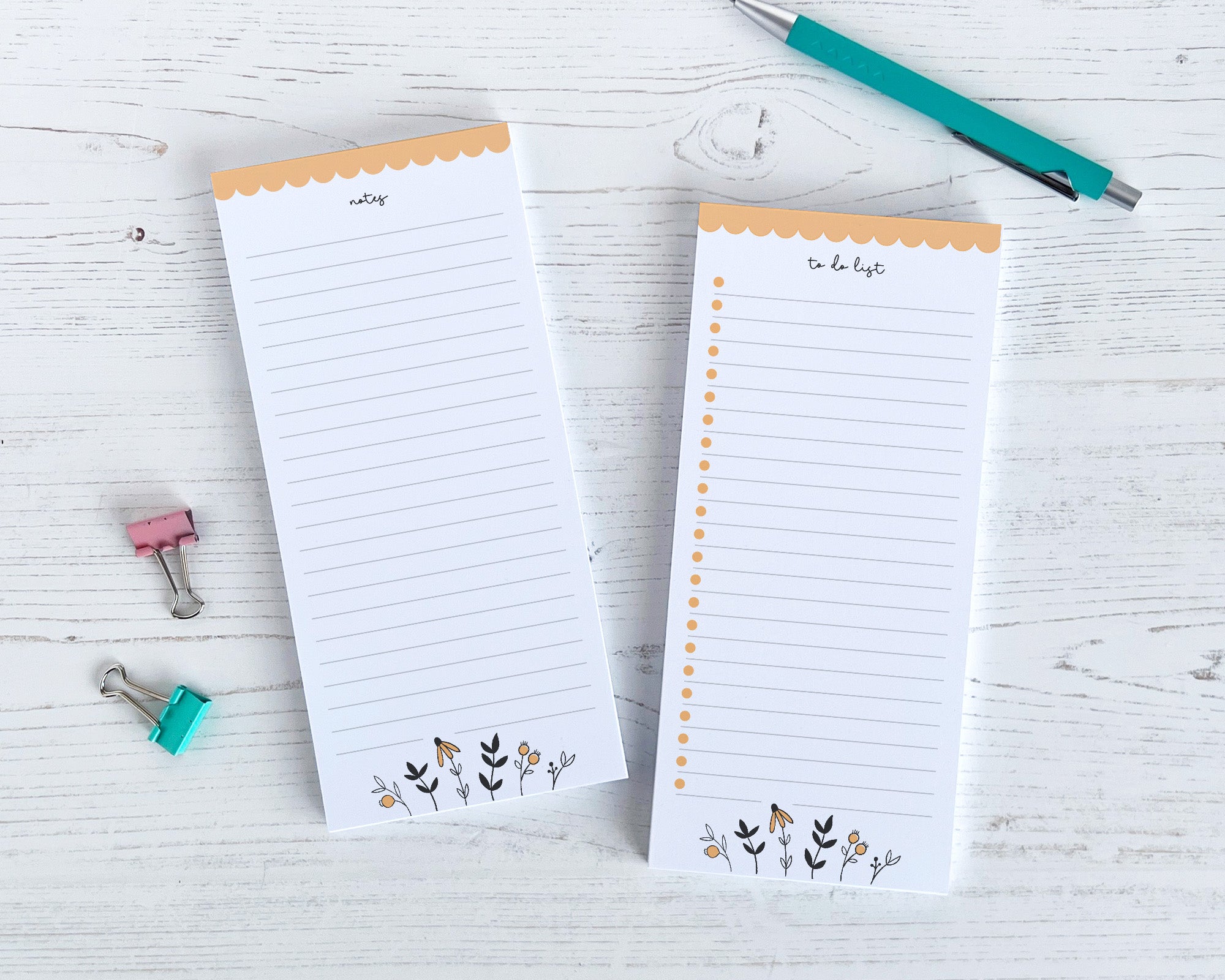 Two notepads with decorative edges on a light wooden surface with a pen and clips.