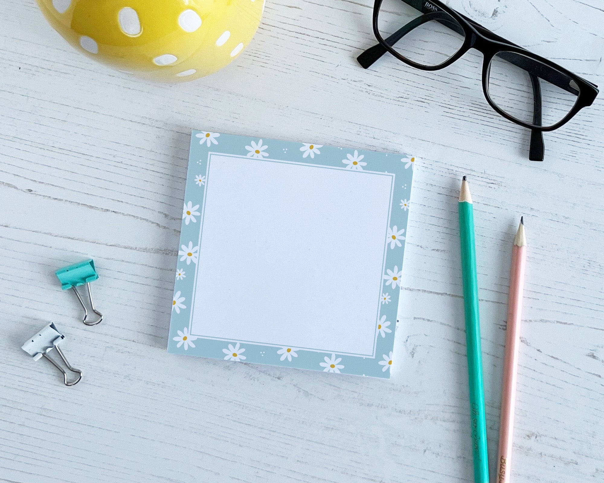 Notepad with daisy design, glasses, pencils, and paper clips on a light surface