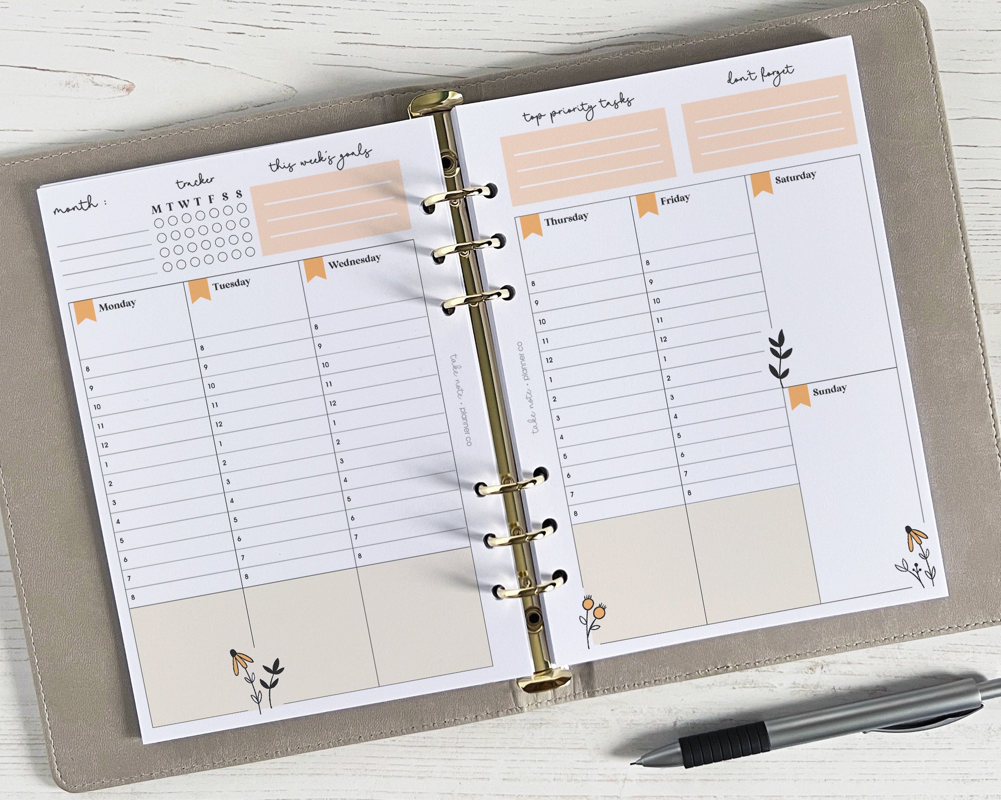 A floral design undated weekly planner insert with a golden spiral binding, placed in a notebook with a pen and some decorative leaves on a white wooden surface.