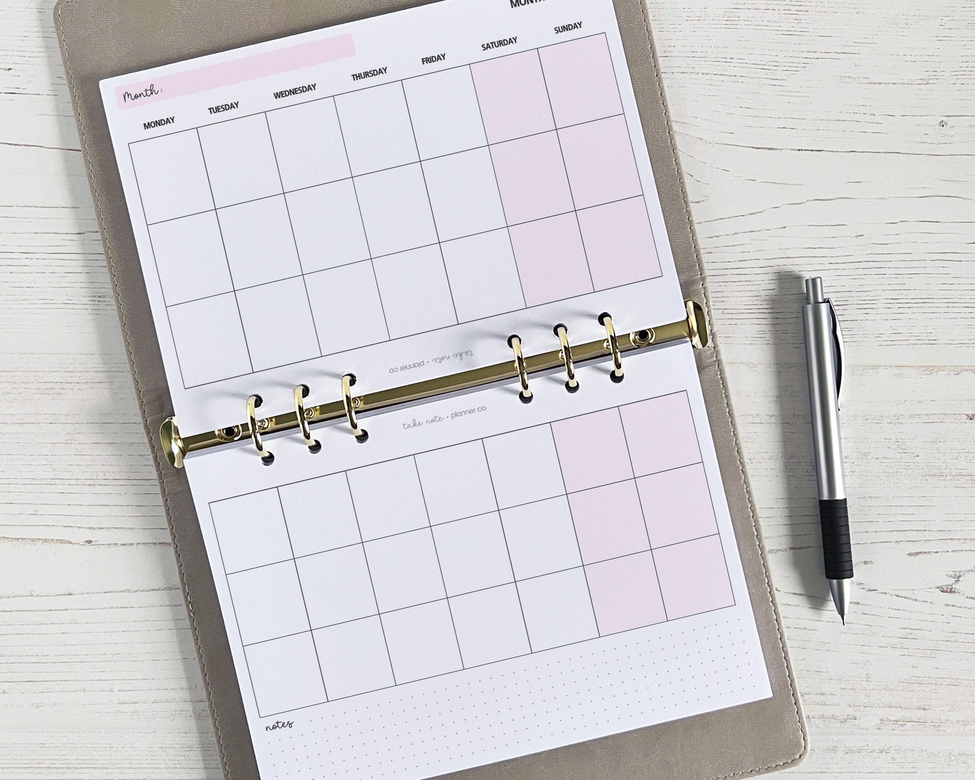 An open monthly planner insert with a landscape layout, featuring boxes for the month ahead and a section for notes or habit tracking, displayed on a wooden surface with a pen beside it.