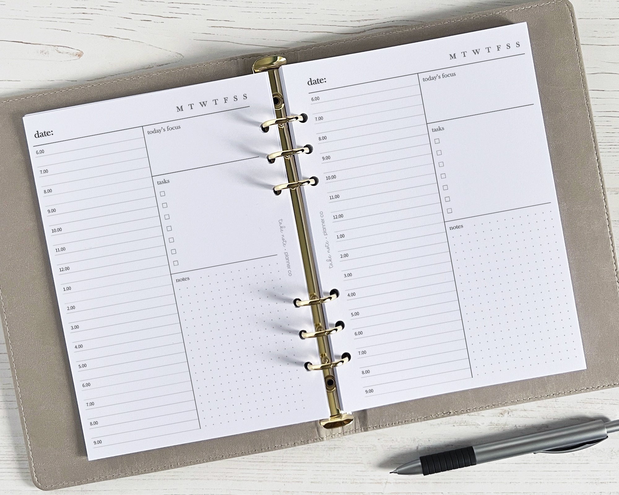 An open daily planner showing undated daily inserts with a simple minimal design, including space for notes, priorities/events, and a to do list. The planner is lying flat on a surface with a pen beside it.