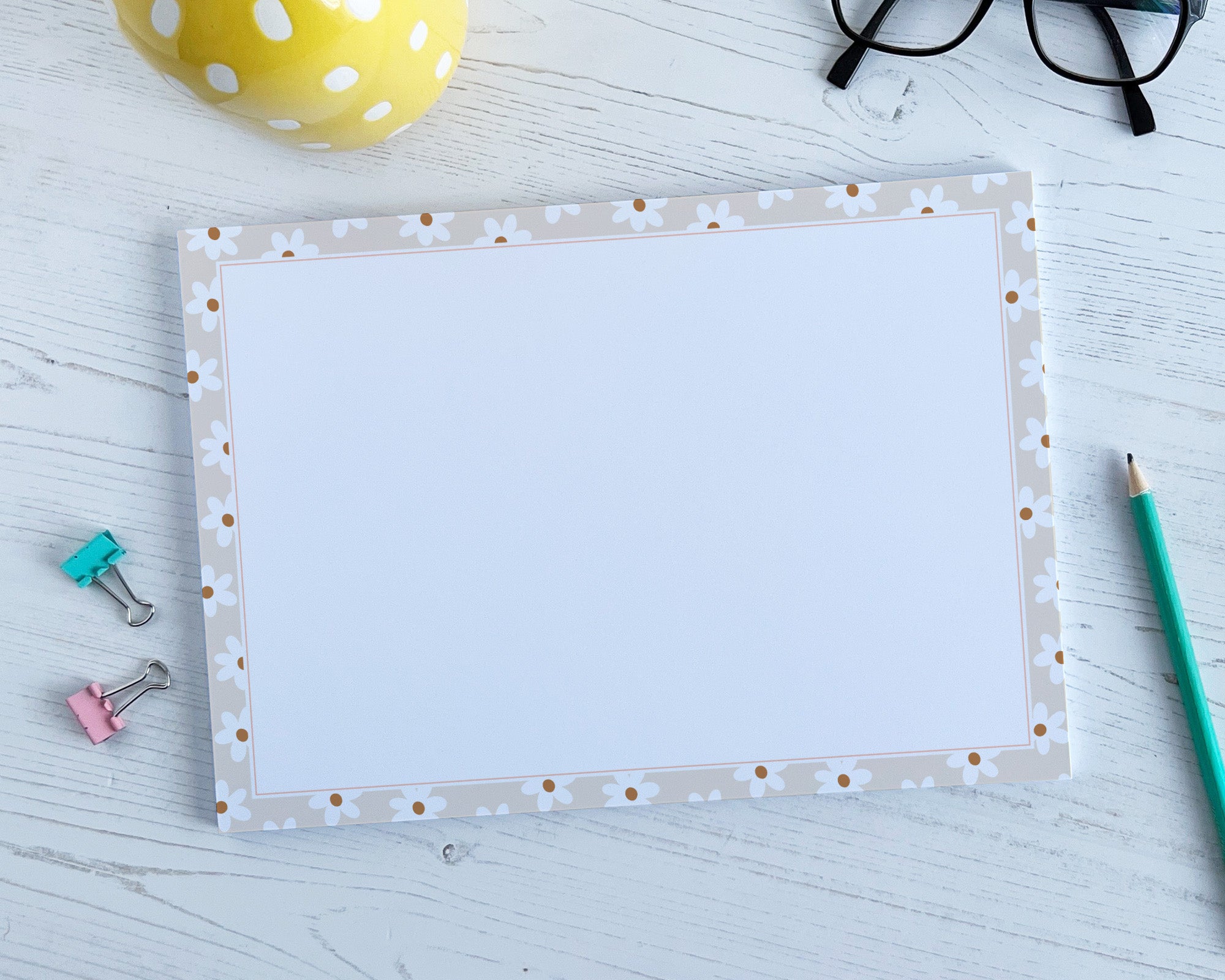 A4 deskpad with decorative  daisy border on a light wooden surface with stationery items.