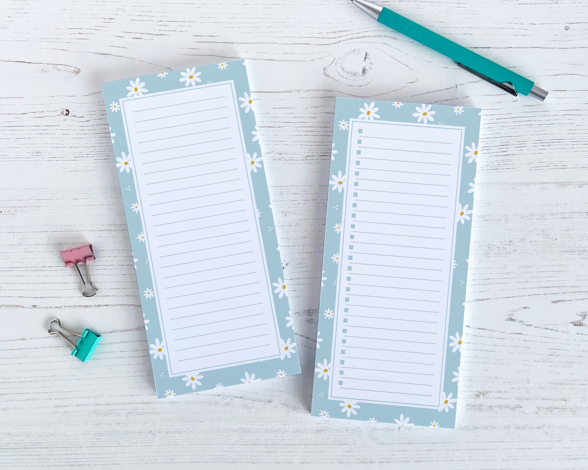 Two floral-themed notepads with a pen and clips on a light wooden surface