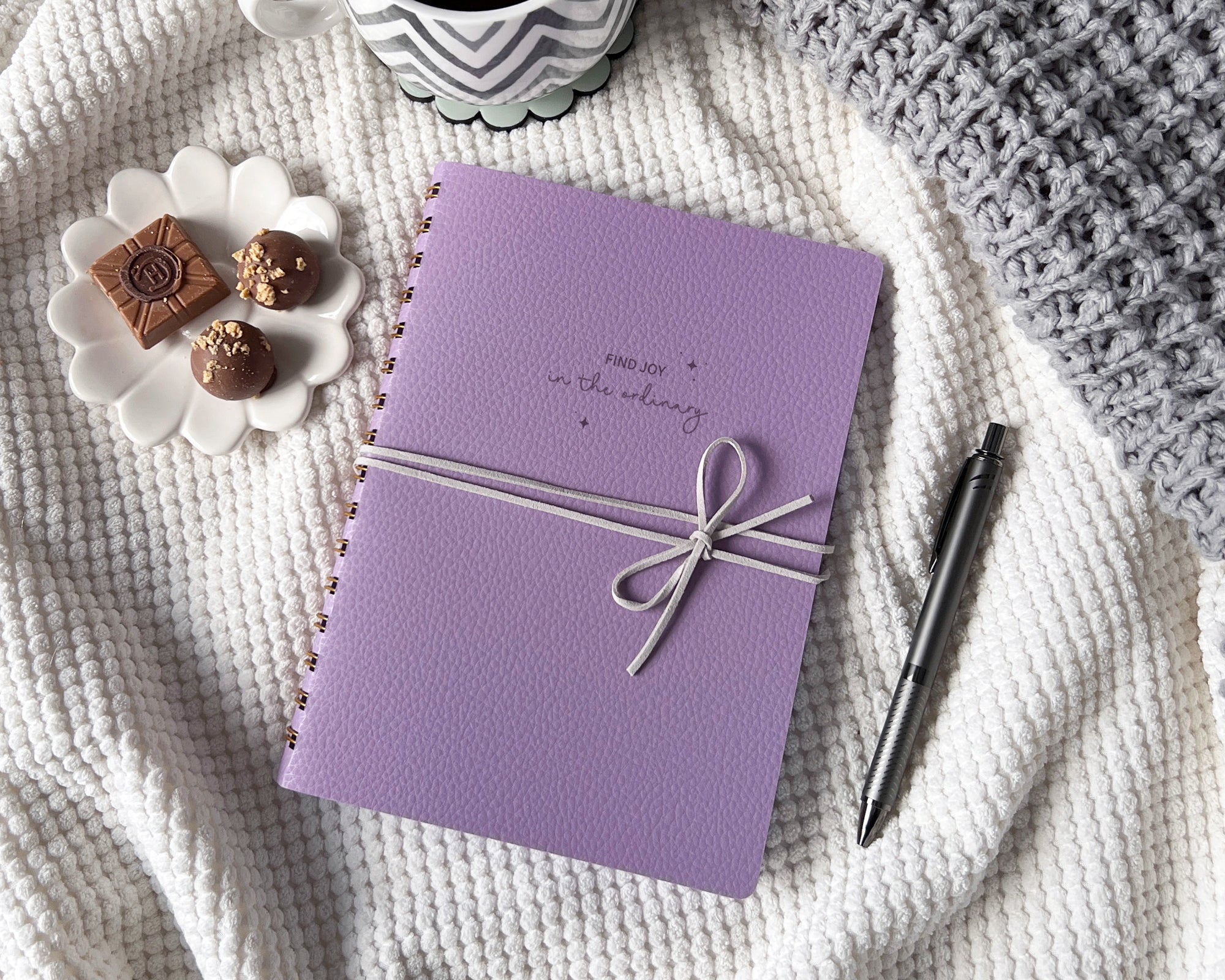 Lavender notebook with a suede cord, pen, and small snacks on a blanket.