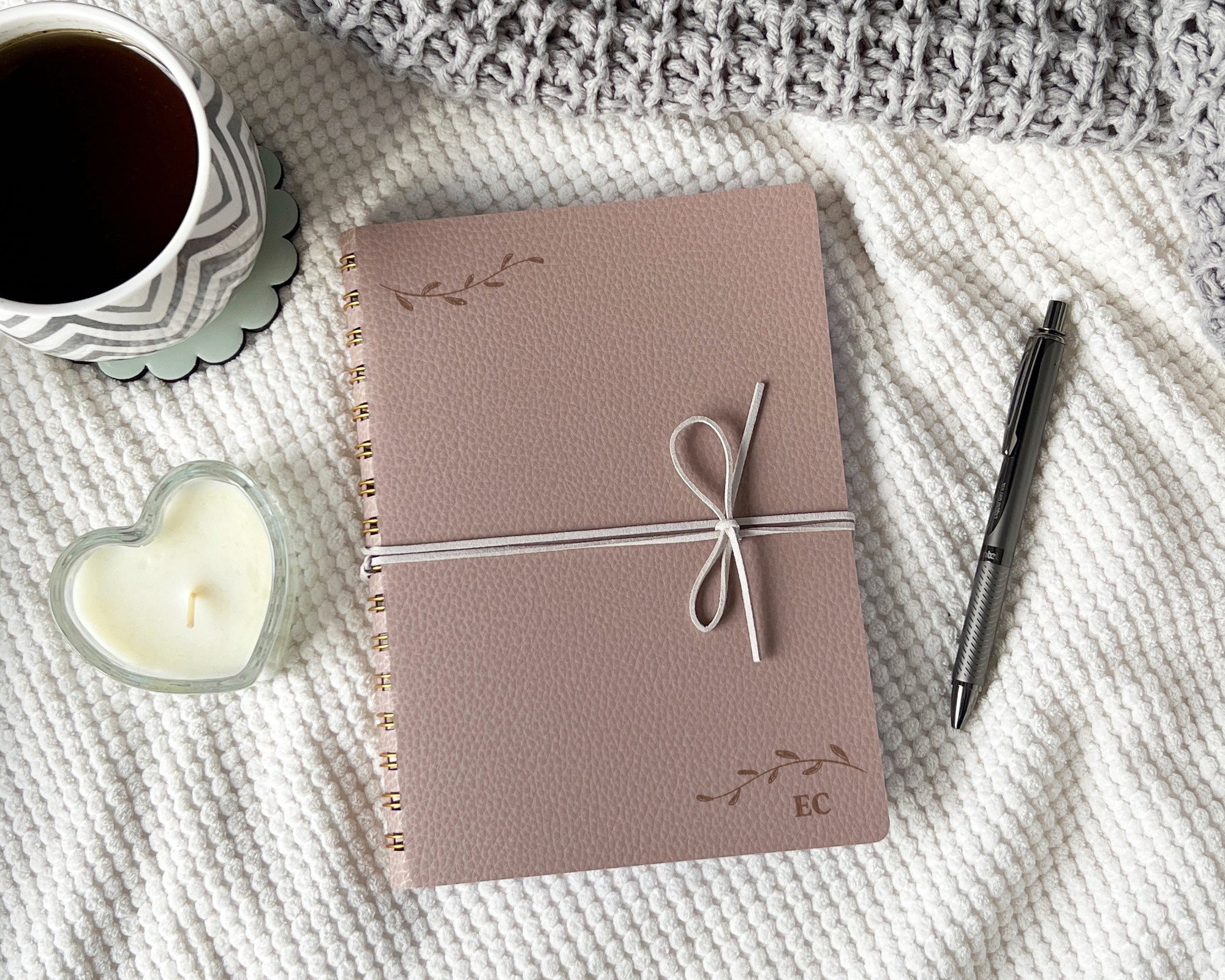Taupe journal tied with a suede cord on a blanket with a cup of coffee, pen, and candle.
