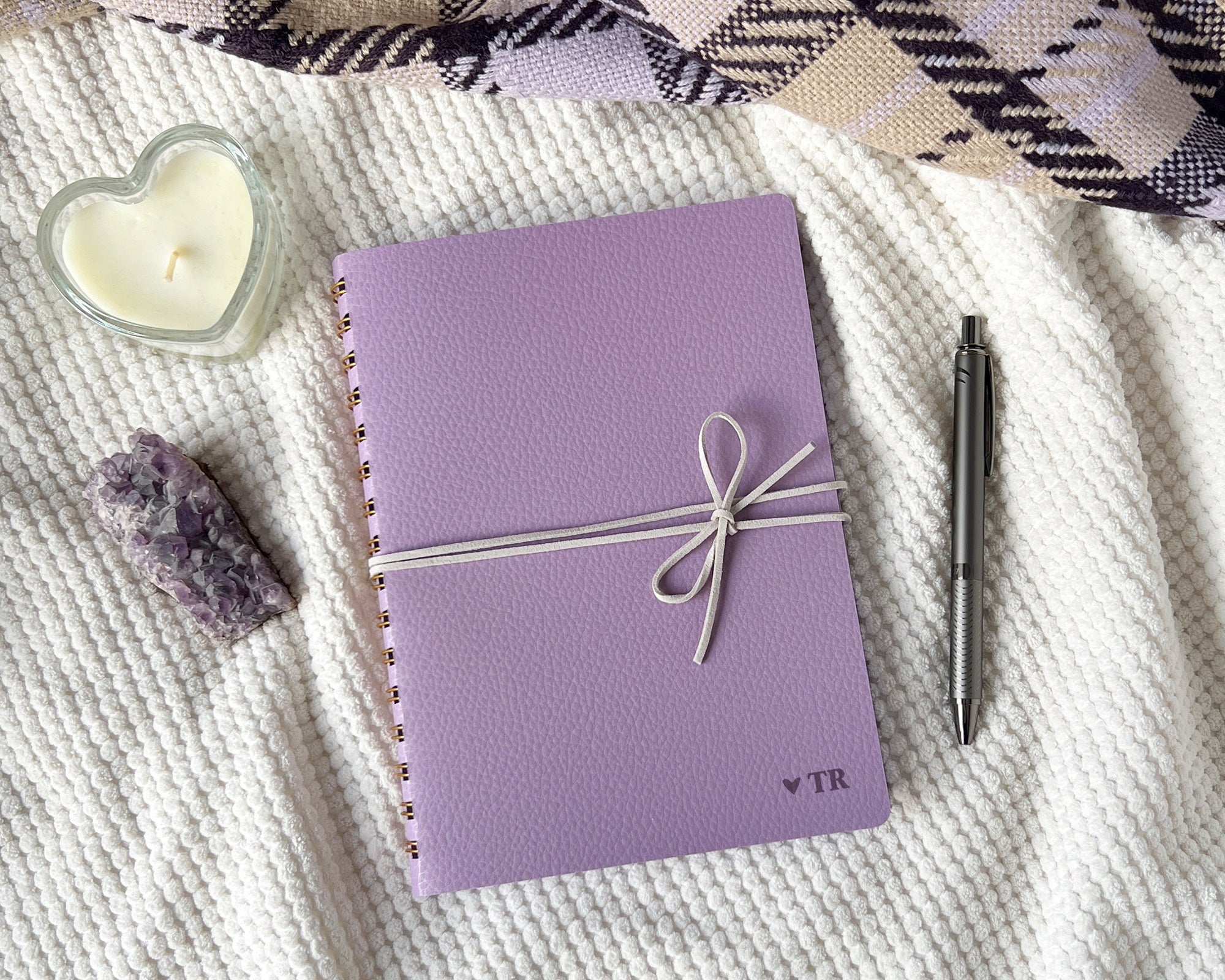 Purple notebook tied with a suede cord, next to a pen and heart-shaped candle on a blanket.